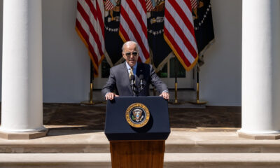 Biden, wearing sunglasses, standing at podium with three American and three Presidential flags hanging behind him.