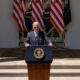 Biden, wearing sunglasses, standing at podium with three American and three Presidential flags hanging behind him.