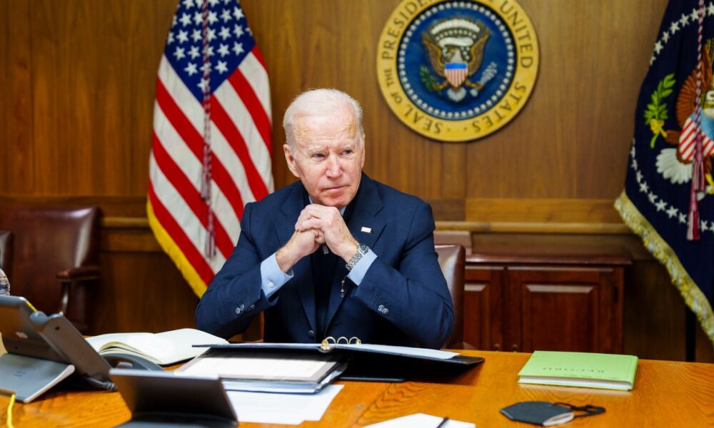 Joe Biden at desk or Cabinet Room table with paneled wall with Presidential Seal behind him.