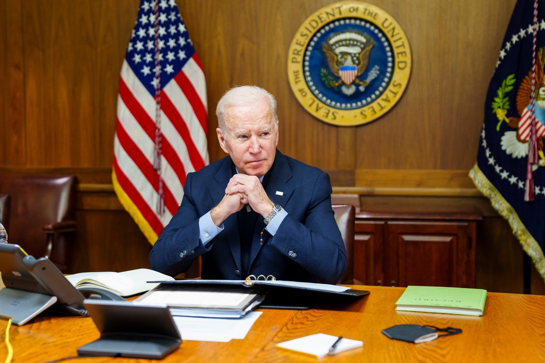 Joe Biden at desk or Cabinet Room table with paneled wall with Presidential Seal behind him.