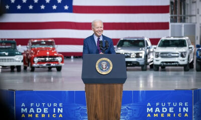Joe Biden poses in front of a stage with ostensible American-made models, maybe some EV models, behind him.