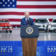 Joe Biden poses in front of a stage with ostensible American-made models, maybe some EV models, behind him.