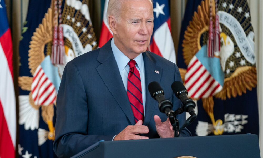 Biden in right front angle, at Presidential podium, with American and Presidential flags on posts behind him