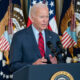 Biden in right front angle, at Presidential podium, with American and Presidential flags on posts behind him