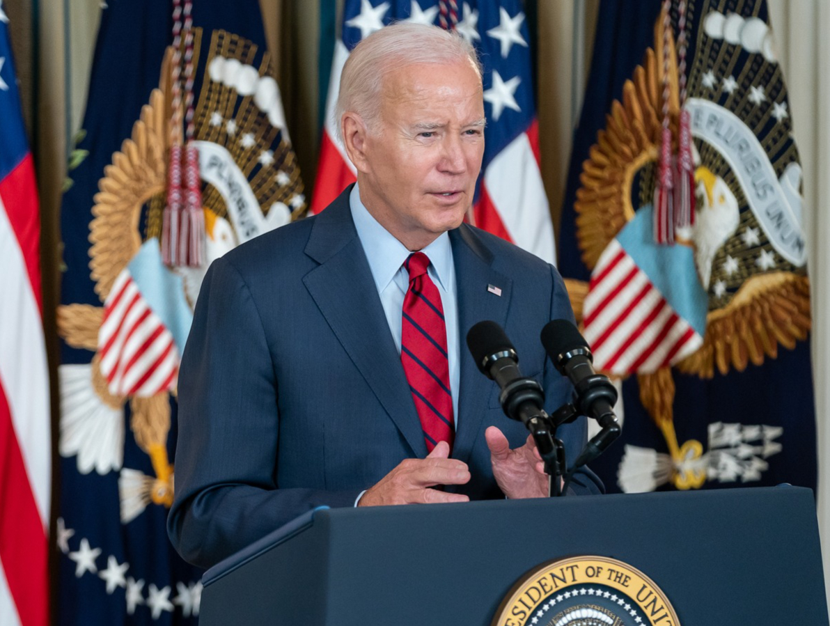 Biden in right front angle, at Presidential podium, with American and Presidential flags on posts behind him