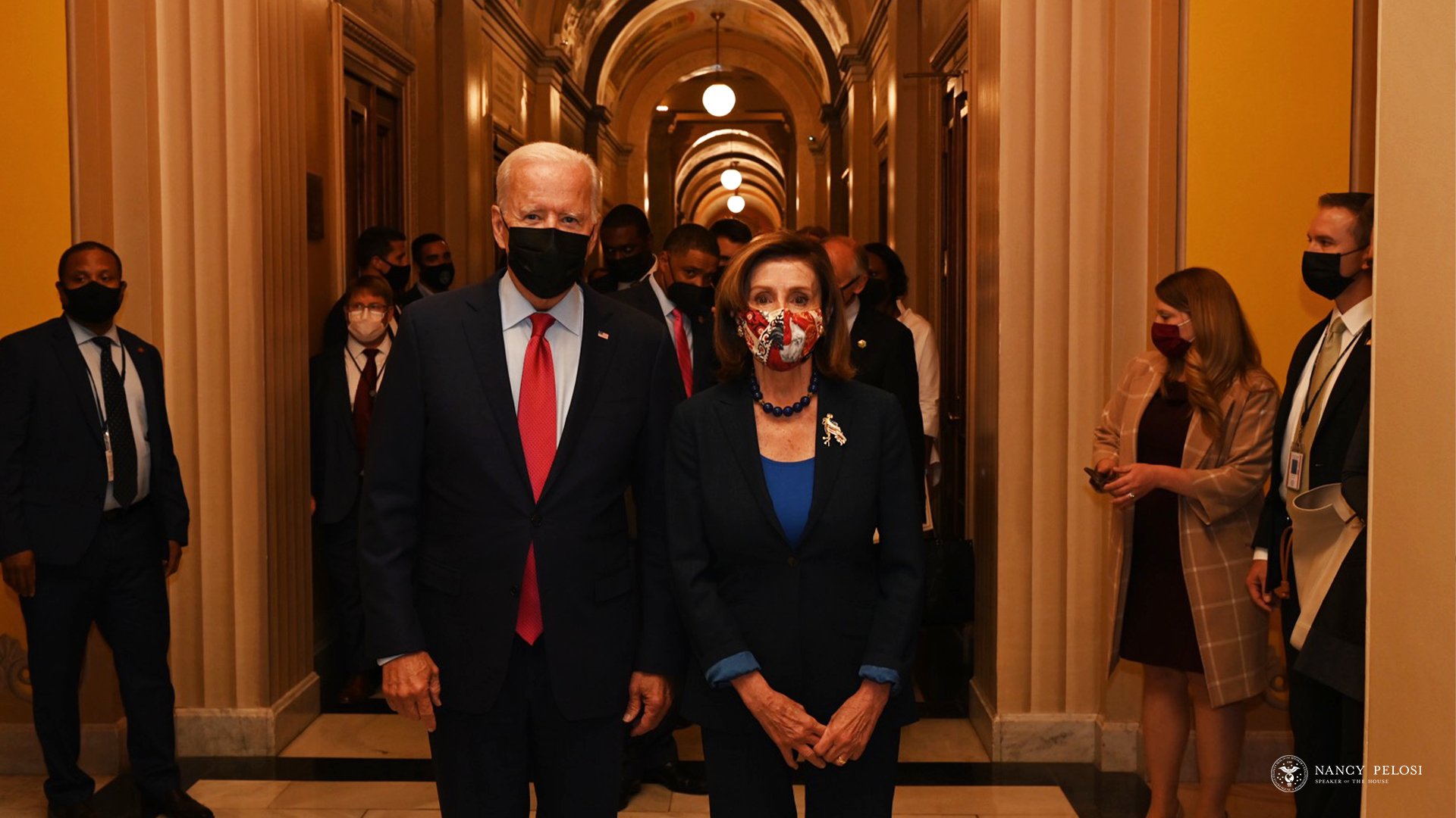 Joe Biden and Nancy Pelosi walk down a corridor - and everyone is masked up in fear of the dreaded coronavirus, putative cause of COVID