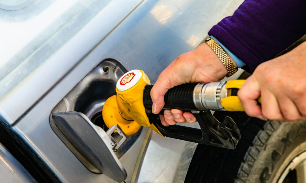 A man holds a gasoline pump nozzle. The stylized seashell, yellow with red outline, is a registered trademark of Royal Dutch Shell. Photo by Jesse Donoghue on Unsplash.com.