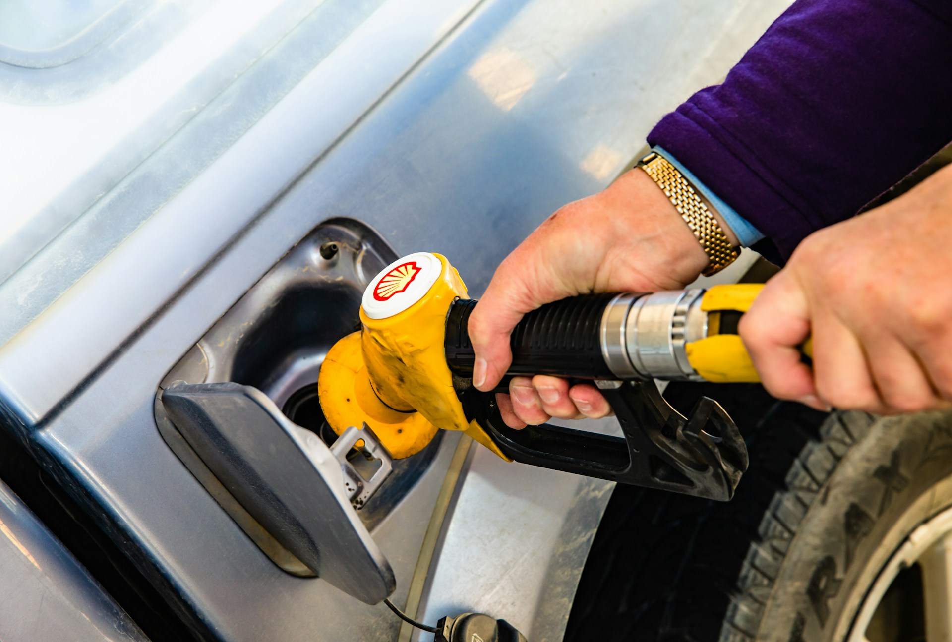 A man holds a gasoline pump nozzle. The stylized seashell, yellow with red outline, is a registered trademark of Royal Dutch Shell. Photo by Jesse Donoghue on Unsplash.com.